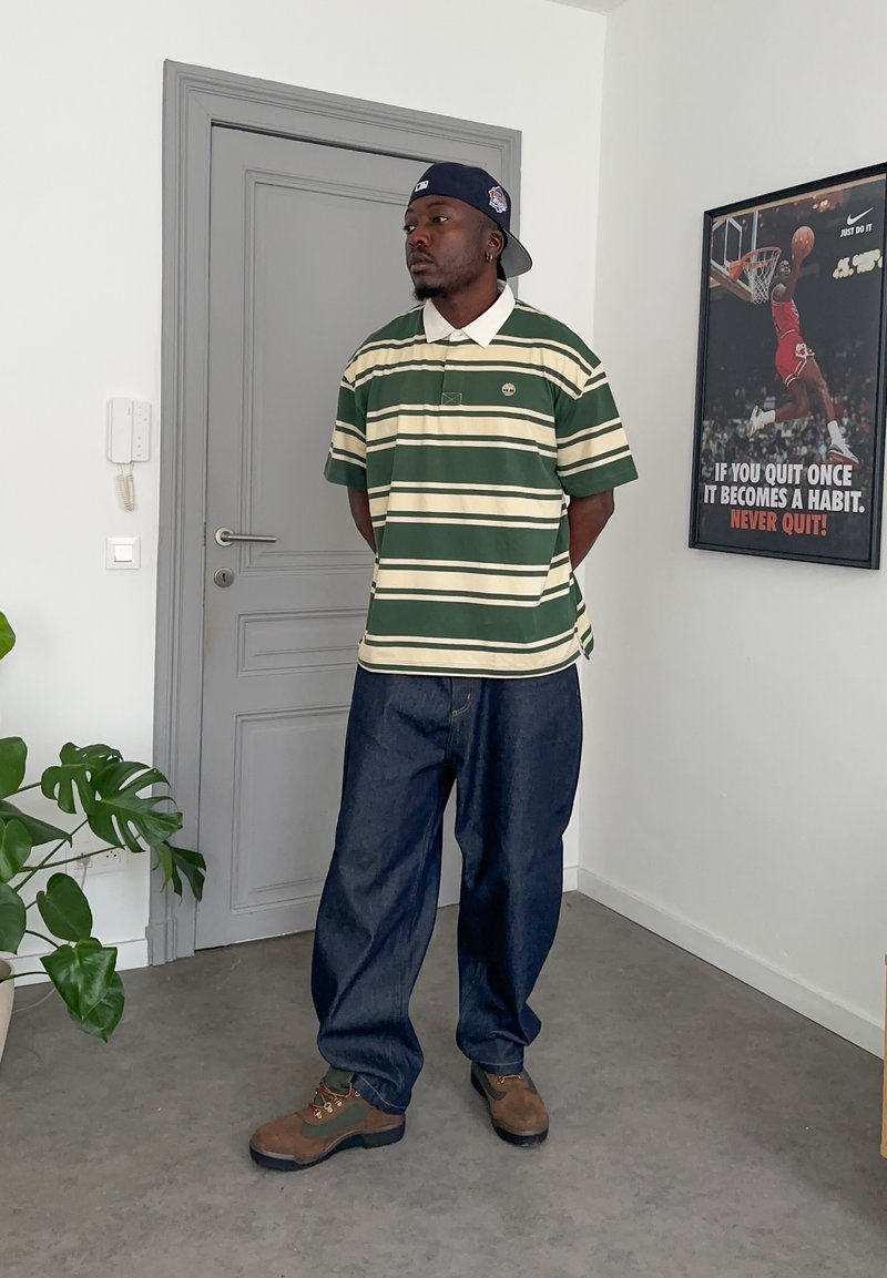 Mann trägt ein gestreiftes grün-beiges Hemd, dunkle lockere Jeans, braune Stiefel und eine rückwärts getragene Kappe, steht in einem Raum mit einem motivierenden Basketball-Poster.