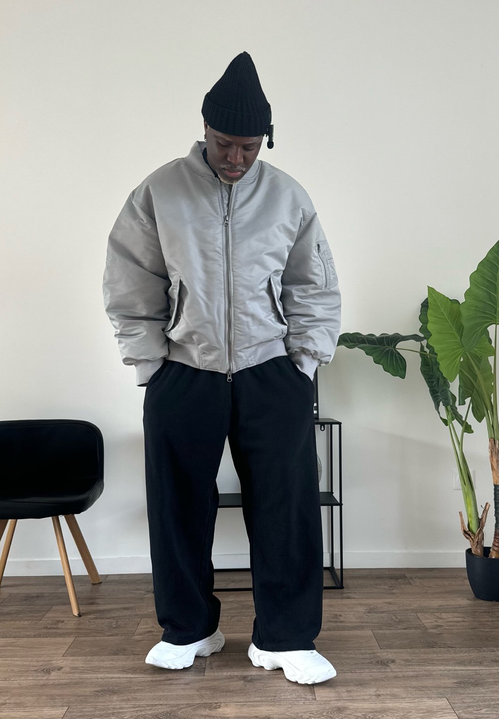 Veste bomber grise avec une fermeture éclair à l'avant, bonnet noir, pantalon large noir et baskets blanches, dans un intérieur minimaliste avec une plante.