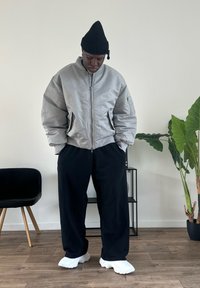Veste bomber grise avec une fermeture éclair à l'avant, bonnet noir, pantalon large noir et baskets blanches, dans un intérieur minimaliste avec une plante.