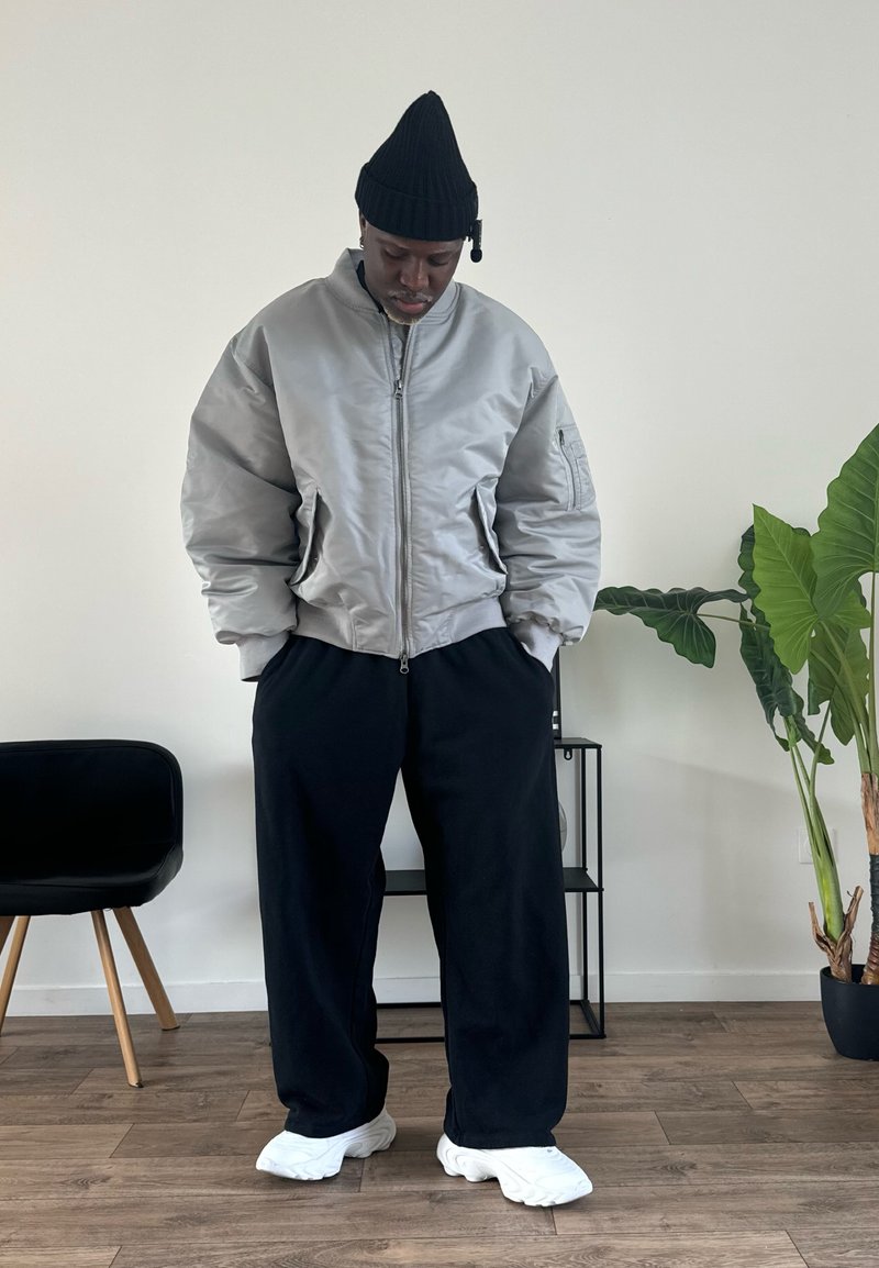 Grey bomber jacket with a zip front, black beanie, black wide-leg pants, and white sneakers, set in a minimal interior with a plant.