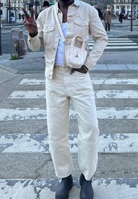 Chaqueta de mezclilla beige y pantalones, combinados con botas negras hasta el tobillo. Un pequeño bolso cruzado beige con un acento de logo. De pie en un paso de cebra.