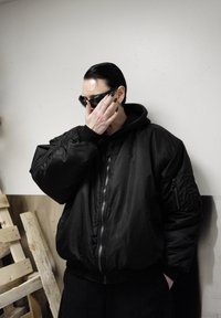 Veste bomber noire avec capuche, fermeture éclair et poches. Portée avec des lunettes de soleil ; tissu texturé, coupe ample et poignets élastiques.