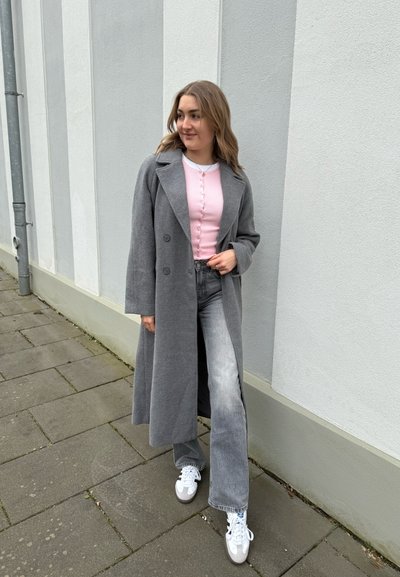 Mujer de pie en la acera, vistiendo un abrigo gris largo, un suéter rosa con botones, jeans grises y zapatillas blancas, mirando hacia un lado.