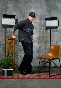 Jeune homme portant une casquette noire et une tenue noire posant entre deux projecteurs de studio, debout à côté d'un grand présentoir en bois et d'une chaise marron sur un tapis à motifs.