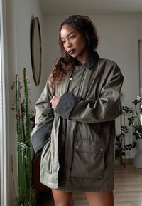 Femme aux longs cheveux tressés portant une veste oversize vert olive, debout à l'intérieur près de grands cactus à côté d'une fenêtre.