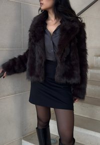 Donna che indossa un cappotto di pelliccia scuro sopra una blusa scura, una mini gonna nera, collant trasparenti e stivali neri fino al ginocchio, in piedi su scale di pietra chiara.
