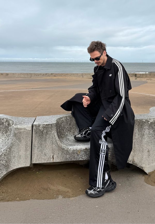 Un homme vêtu d’une tenue de sport noire à rayures et portant des lunettes de soleil est assis sur un mur en béton au bord de la mer et regarde vers le bas près du rivage.