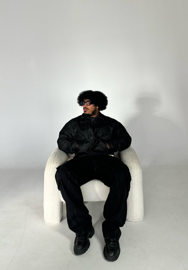 Zwarte denimjasje, losse zwarte broek en stevige zwarte schoenen gedragen door een persoon die zit in een witte, gebogen textuurstoel tegen een lichte achtergrond.