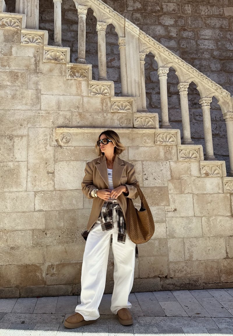 Femme en blazer oversize, pantalon blanc et lunettes de soleil, se tient devant un mur en pierre historique avec un escalier orné, sous un jour lumineux.