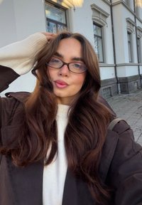 Brun ytterjakke med en kremfarget innergenser, langt bølget mørkt hår, firkantede briller og en lys bakgrunn med en bygning.