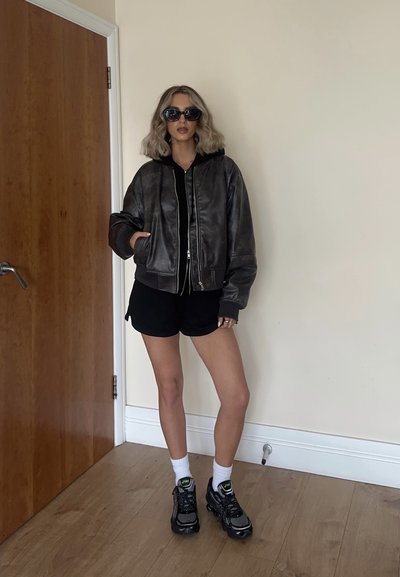 Chaqueta bomber de cuero negra con capucha, combinada con una camiseta negra y pantalones cortos. Zapatillas oscuras, calcetines blancos; de pie contra una pared clara.