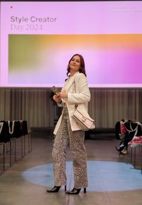 Bílý přizpůsobený blejzr, široké kalhoty se zvířecím potiskem, černé podpatky a světle růžová kabelka se zlatými detaily, na pódiu s barevným pozadím.