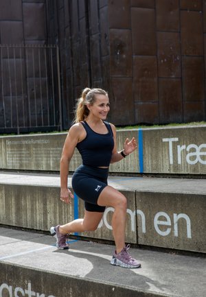 Top deportivo negro sin mangas y pantalones cortos; zapatillas de correr moradas claras. La persona está subiendo una escalera de hormigón con marcas y texto azules.