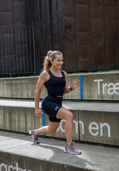 Top deportivo negro sin mangas y pantalones cortos; zapatillas de correr moradas claras. La persona está subiendo una escalera de hormigón con marcas y texto azules.