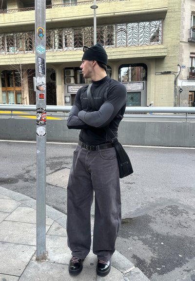 Joven con gorro negro, camiseta ajustada de manga larga, pantalones anchos grises y zapatos negros, de pie con los brazos cruzados en la acera de la ciudad.