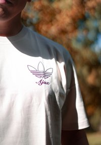 Tricou din bumbac de culoare crem, cu un logo trefoil Adidas de culoare violet și textul "Ajax" dedesubt, cu mâneci scurte și decolteu rotund.