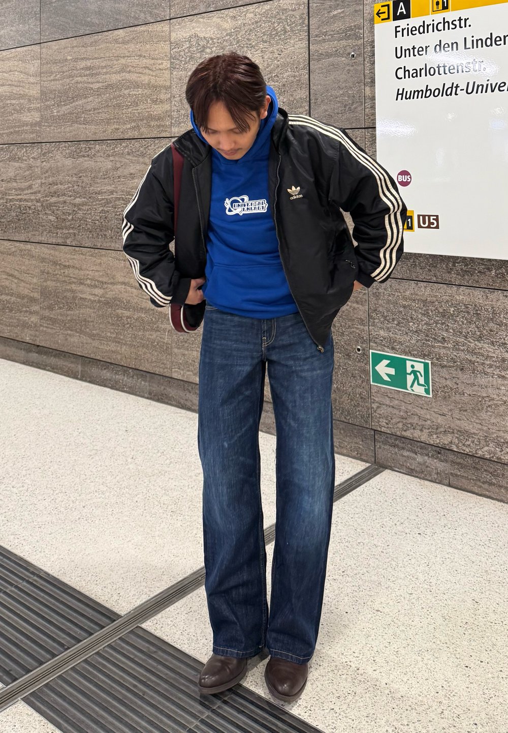Veste de sport noire avec bandes blanches, sweat à capuche bleu avec logo, jean bleu, chaussures marron, debout dans un environnement intérieur avec un mur carrelé.