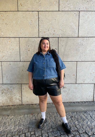 Camisa de mezclilla azul de manga corta con rayas verticales, pantalones cortos de cuero negros, calcetines blancos y zapatos negros de plataforma; de pie contra una pared texturizada.