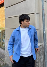 Camisa azul claro abotonada sobre una camiseta blanca, combinada con pantalones negros; tela suave con detalles de botones y un corte relajado.