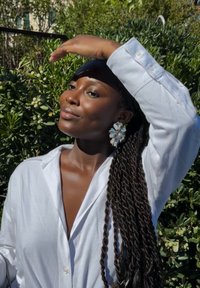 Camicia bianca con bottoni, capelli intrecciati, grandi orecchini floreali blu e bianchi. Mano alzata, con vegetazione sullo sfondo. Texture della pelle liscia.