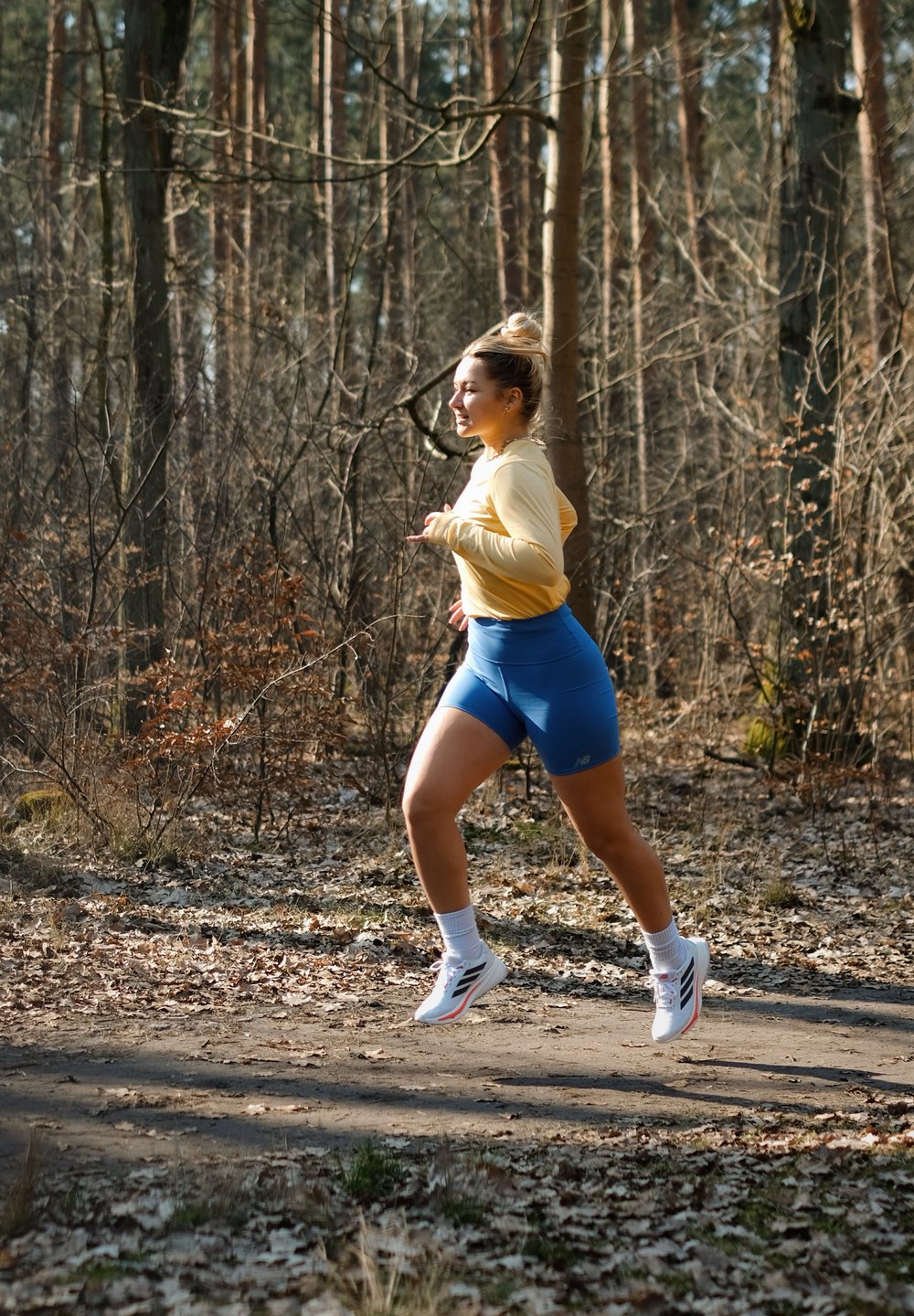 Corredor com uma camisola de manga longa amarela, calções azuis e ténis brancos, a correr por um caminho na floresta com árvores e folhas secas ao fundo.