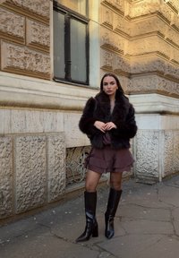Femme portant une veste en fourrure sombre, une jupe marron à volants, des bottes noires montantes, debout devant un bâtiment en pierre avec une fenêtre décorative.