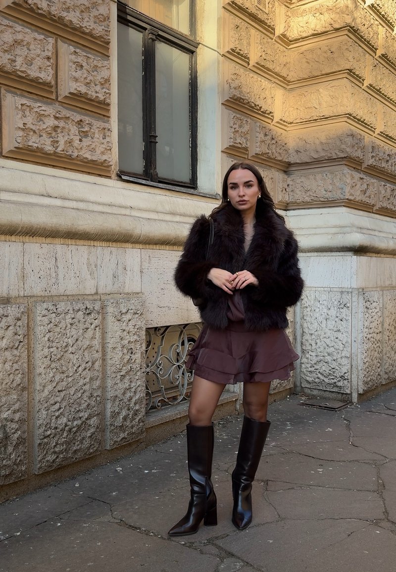 Femme portant une veste en fourrure sombre, une jupe marron à volants, des bottes noires montantes, debout devant un bâtiment en pierre avec une fenêtre décorative.