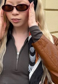 Hnědá kožená bunda přes žebrovaný tmavě šedý top, s tmavými oválnými slunečními brýlemi a s šátkem s vzorem kolem krku.