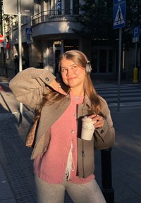 Jonge vrouw in een roze trui en een grijze jas houdt een koffiemok vast, draagt een koptelefoon en poseert lachend op een zonnige stadsstraathoek.