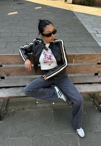 Veste en cuir noir avec des rayures blanches, t-shirt graphique blanc, jeans foncés et baskets blanches avec des rayures noires ; assis sur un banc en bois.