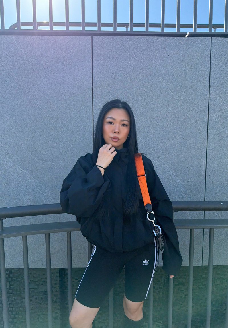 Jeune femme portant un short et une veste Adidas noirs avec un sac à bandoulière orange, debout près d'une rambarde métallique et d'un mur carrelé gris en extérieur.