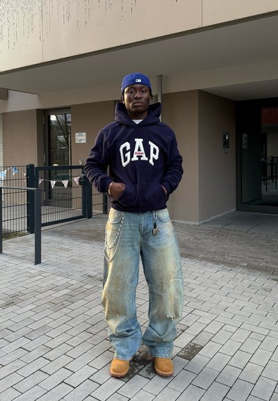 Sudadera de forro polar de la Marina con el logo "GAP", jeans desgastados de color azul claro, botas marrones y detalle de cadena. El fondo presenta la fachada de un edificio moderno.