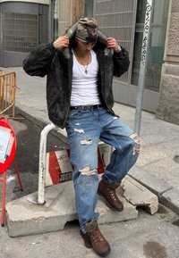 Črna usnjena jakna z ovratnikom iz krzna, bela majica brez rokavov, strgane modre kavbojke in rjavi pohodniški škornji. Mestno okolje z gradbenimi ovirami.