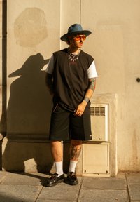 Übergroßes schwarzes ärmelloses Shirt über einem weißen T-Shirt, schwarze Shorts, bunte Perlenkette, breiter blauer Hut, orangefarbene Sonnenbrille, schwarze Schuhe.