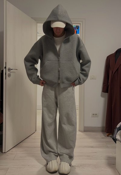 Persona que lleva una sudadera gris con cremallera de gran tamaño, pantalones de chándal anchos a juego, una gorra blanca y zapatillas blancas sin cordones, de pie en el interior.