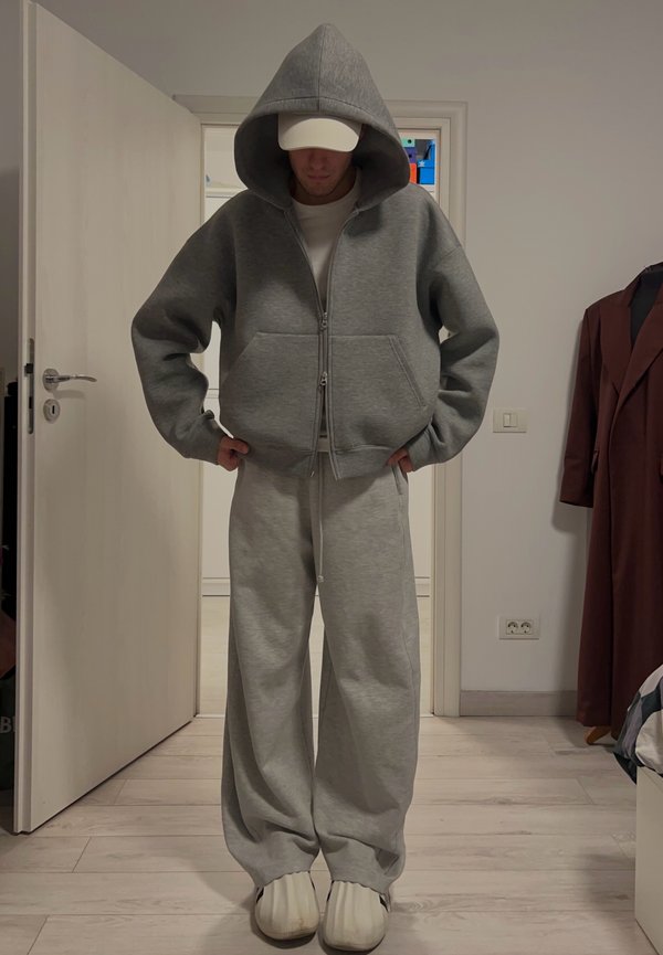 Person iført en overdimensjonert grå hettegenser med glidelås, matchende vide joggebukser, hvit caps og hvite slip-on sko som står innendørs.