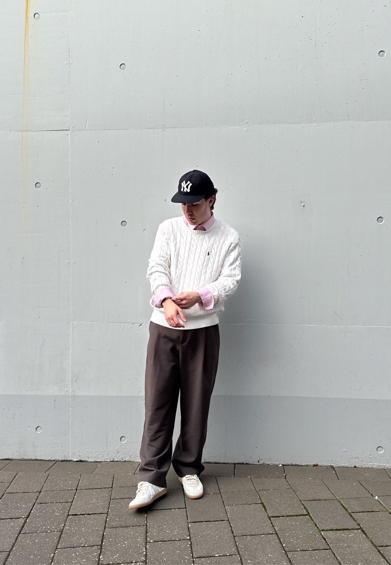 Jeune homme ajustant la manche de son pull, portant un pull torsadé blanc, une chemise rose, un pantalon foncé, des baskets blanches et une casquette de baseball noire, debout sur un pavé carrelé.
