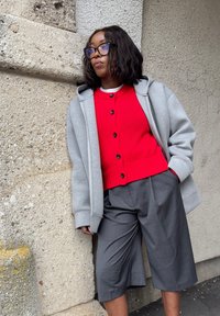 Felpa grigia oversize, maglione rosso a coste con bottoni e culotte grigie sartoriali. L'insieme presenta texture e colori a contrasto.