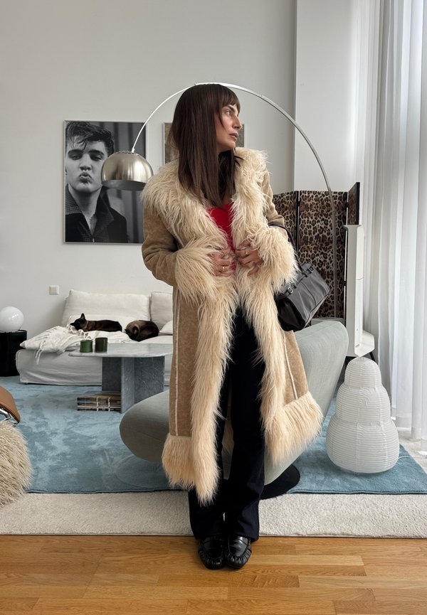 Une femme vêtue d’un long manteau beige à bordure en fourrure crème pose dans un salon moderne, en maintenant son manteau fermé ; derrière elle, deux chats se reposent sur un canapé.