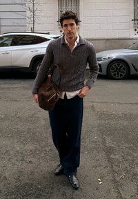 Jeune homme portant un pull gris en maille torsadée, un pantalon bleu marine et des chaussures noires, marchant dans la rue avec un grand sac en cuir marron sur l'épaule.