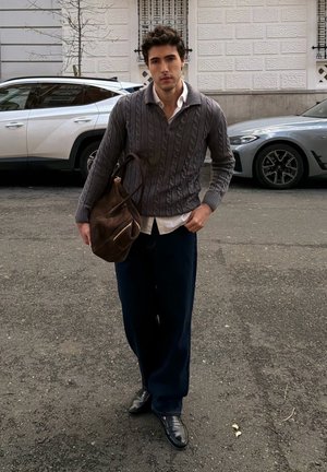 Joven con suéter gris de punto trenzado, pantalones azul marino y zapatos negros camina por la calle llevando un gran bolso de piel marrón sobre el hombro.