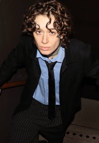 O persoană cu păr creț poartă o jachetă de costum neagră peste o cămașă bleu și o cravată neagră, asortate cu pantaloni cu dungi subțiri. Balustradă de lemn vizibilă.