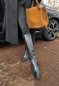 Bottes hautes jusqu'aux genoux en cuir noir avec des bouts pointus et un petit talon. Un sac fourre-tout en daim marron est porté à côté de leggings noirs et d'un manteau.