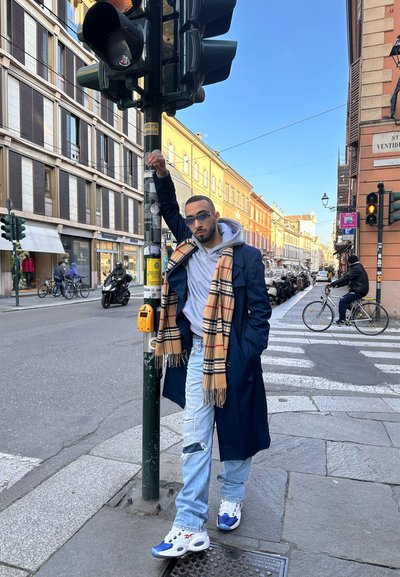 Un joven se apoya en un poste de tráfico, vistiendo un abrigo largo azul marino, sudadera gris, bufanda a cuadros color beige, jeans claros rasgados y zapatillas blancas.