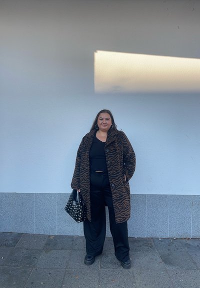 Abrigo a rayas de cebra marrón y negro, top negro, pantalones negros holgados y un bolso negro con tachuelas. De pie contra una pared blanca.