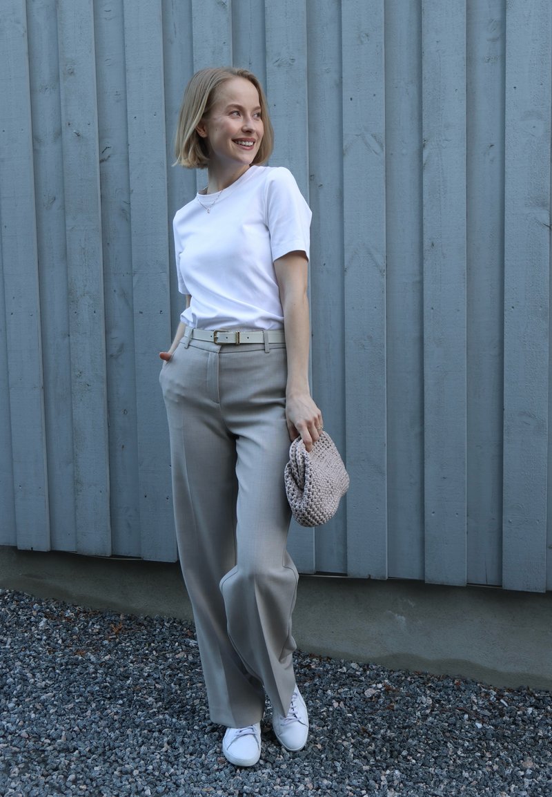 Femme souriante aux cheveux blonds courts se tient à l'extérieur, habillée d'un t-shirt blanc, d'un pantalon gris, de baskets blanches et tenant une pochette beige.