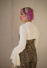 Hvid langærmet bluse med flagrende manchetter, parret med overalls i leopardmønster. Håret er pink med farverige striber. Guldtilbehør til stede.
