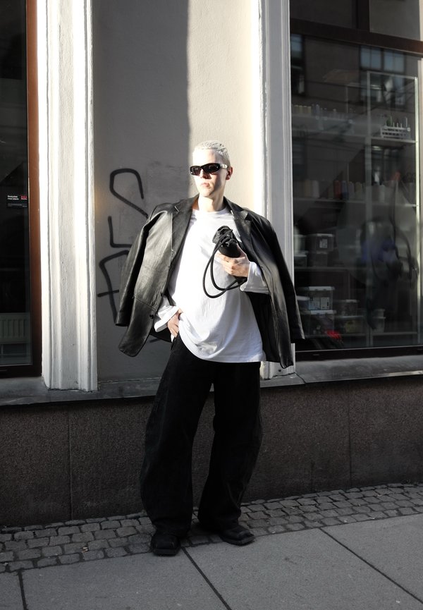 Lühikeste blondide juustega isik, kes kannab musti päikeseprille, üle suuruses musta nahkjakki valge särgi ja mustade laia lõikega pükste peal, seisab kõnniteel.