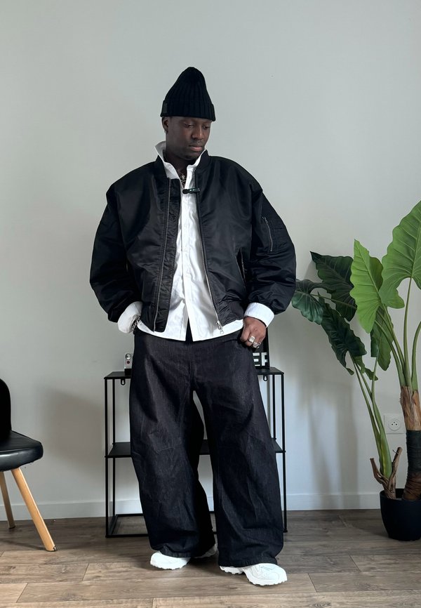 Giacca bomber nera con zip frontale, camicia bianca sotto, pantaloni in denim scuri larghi e sneakers bianche. Foglie di piante circolari sullo sfondo.