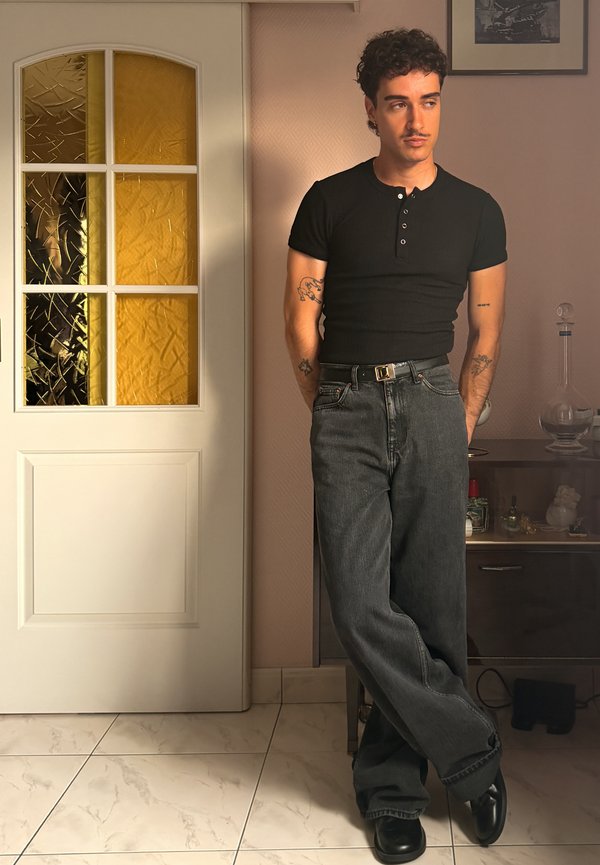 Young man with tattoos wearing black shirt and loose gray jeans stands with legs crossed indoors near a door and cabinet with glassware.